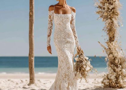Woman in a Boho wedding dress with square neckline and long fitted lace sleeves standing on sandy beach, featuring all-over floral lace pattern and a subtle train, with sleek bun hairstyle and an ocean backdrop with wooden floral arch.