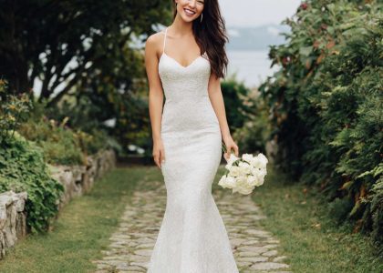 A woman in a shimmering lace Mermaid Tail wedding dress with spaghetti straps stands on a stone path, smiling as she holds a small bouquet of white roses. She wears white pointed-toe shoes, with green trees and an overcast sky in the background.