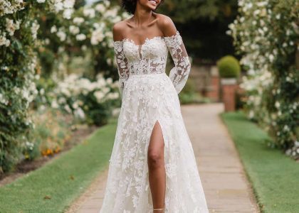Woman in an off-the-shoulder A-Line lace wedding dress with sheer long sleeves and floral lace appliqué, standing on a garden path with greenery and white flowers in the background, wearing strappy sandals and smiling softly.