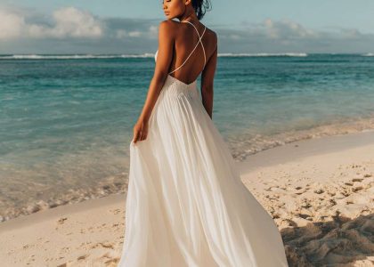 Stunning Black bride in a boho beach wedding dress with A-line silhouette, soft ivory chiffon, deep V-neckline, delicate spaghetti straps, and open back with cross-back detail, standing barefoot on sandy shore during golden hour, with airy train billowing in ocean breeze.