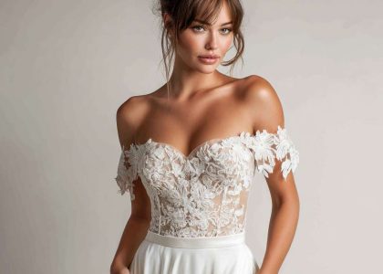 Woman in Boho flowery wedding dress with off-the-shoulder neckline and fitted lace bodice featuring intricate floral embroidery, smooth satin skirt, and loose romantic updo, posing against a neutral background with one hand in pocket.